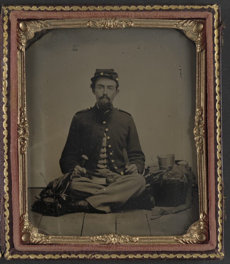An unidentified Union soldier poses for the camera armed with fork, knife, and plate—ready to eat a slice he's just cut from the apple on his lap.