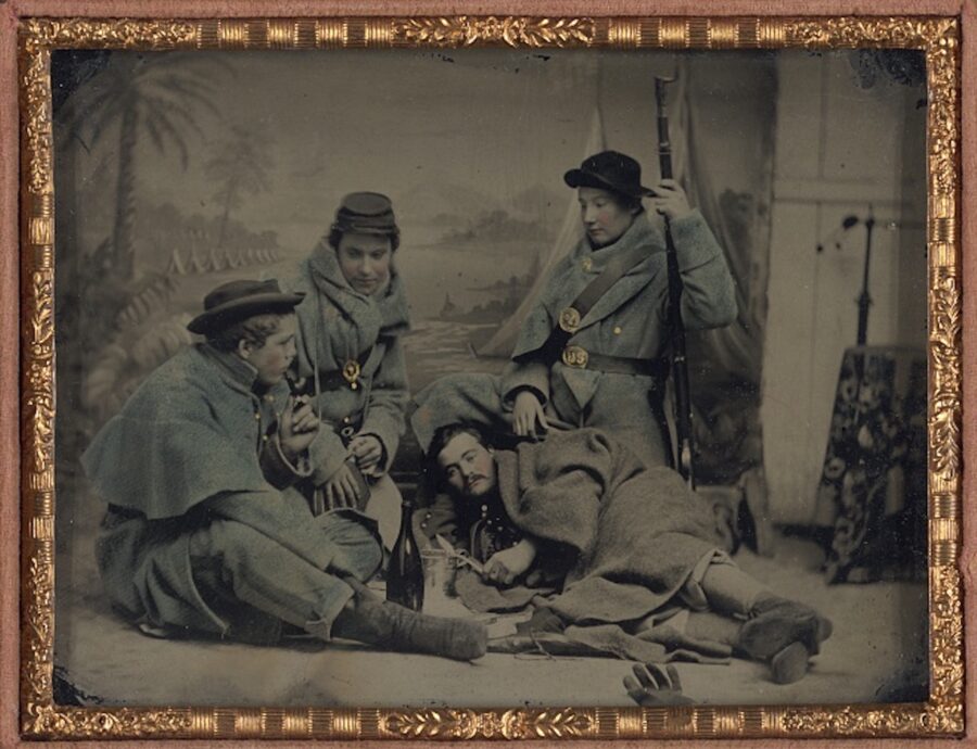 Sergeant Cornelius V. Moore of Company B, 100th New York Volunteers, a sergeant of 39th Illinois Regiment, a corporal of 106th New York Volunteers, and a private of the 11th Vermont Regiment in camp scene poses in front of painted backdrop showing military camp