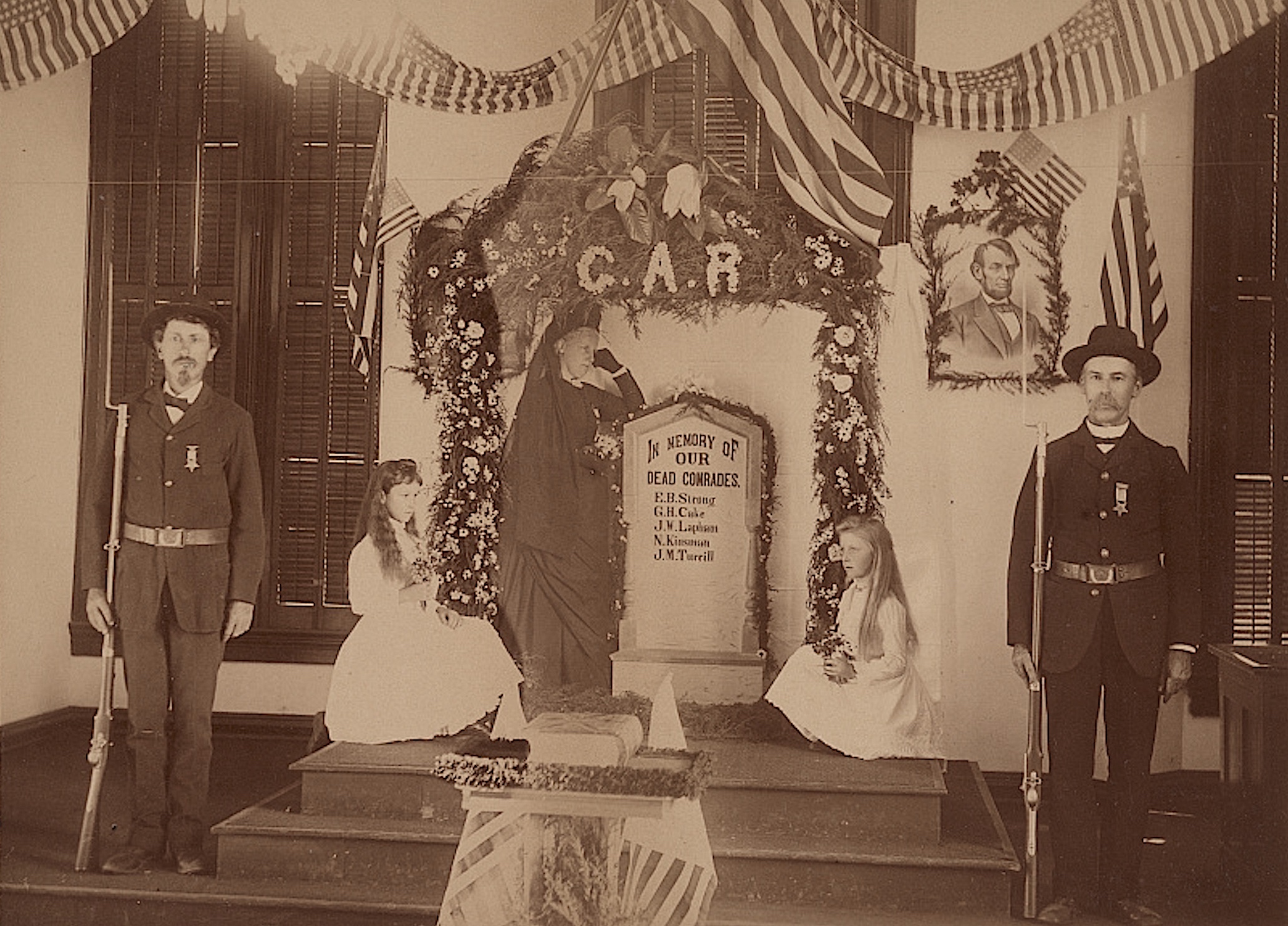 Two Union veterans, members of General John Sedgwick GAR Post #17 of Santa Ana, California, stand guard in front of an altar to dead comrades, a woman in mourning attire and two girls holding flowers behind them. The image was likely made in the late 1800s.