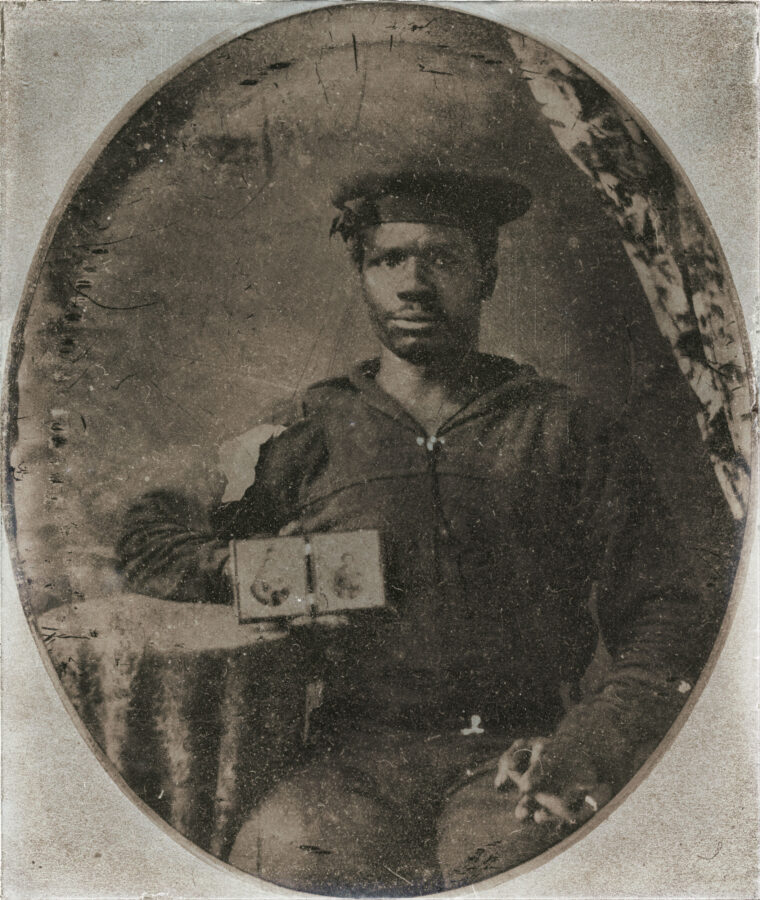 African-American Civil War sailor holds a photo of Confederate soldiers in one hand and a half-smoked cigar in the other.