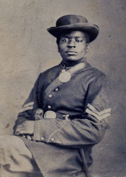 Portrait photograph of Charles Mudd, Corporal, Co. C, 108th U.S. Colored Infantry, seated in uniform with hat